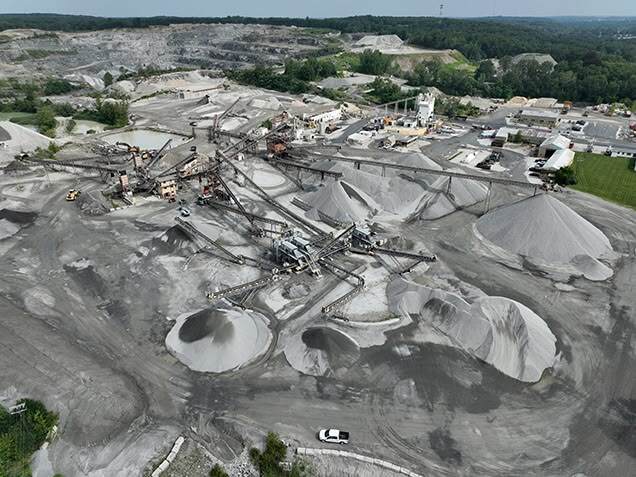 Aerial view of a quarry with conveyor belts, heavy machinery, and large piles of gravel and sand spread across the excavation site