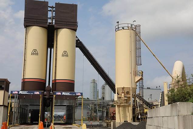 Two tall beige silos with “ASTEC” logos at an asphalt plant, connected by a conveyor belt. Industrial machinery and additional storage structures are visible around them.