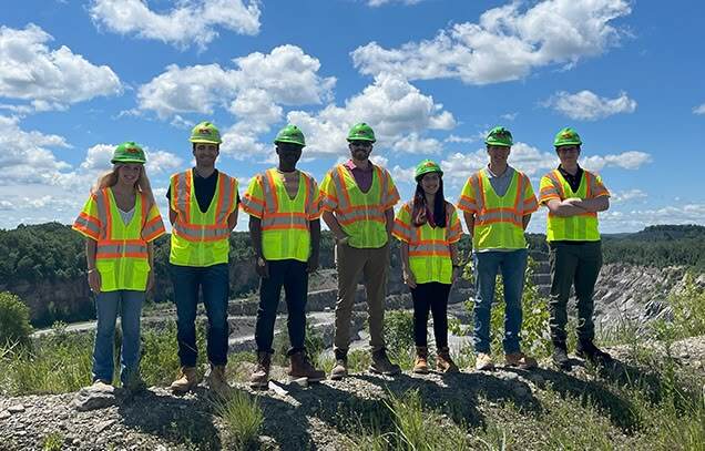  Group of six interns wearing hard hats, safety vests, and boots standing in front of a quarry site on a sunny day with a blue sky and clouds.